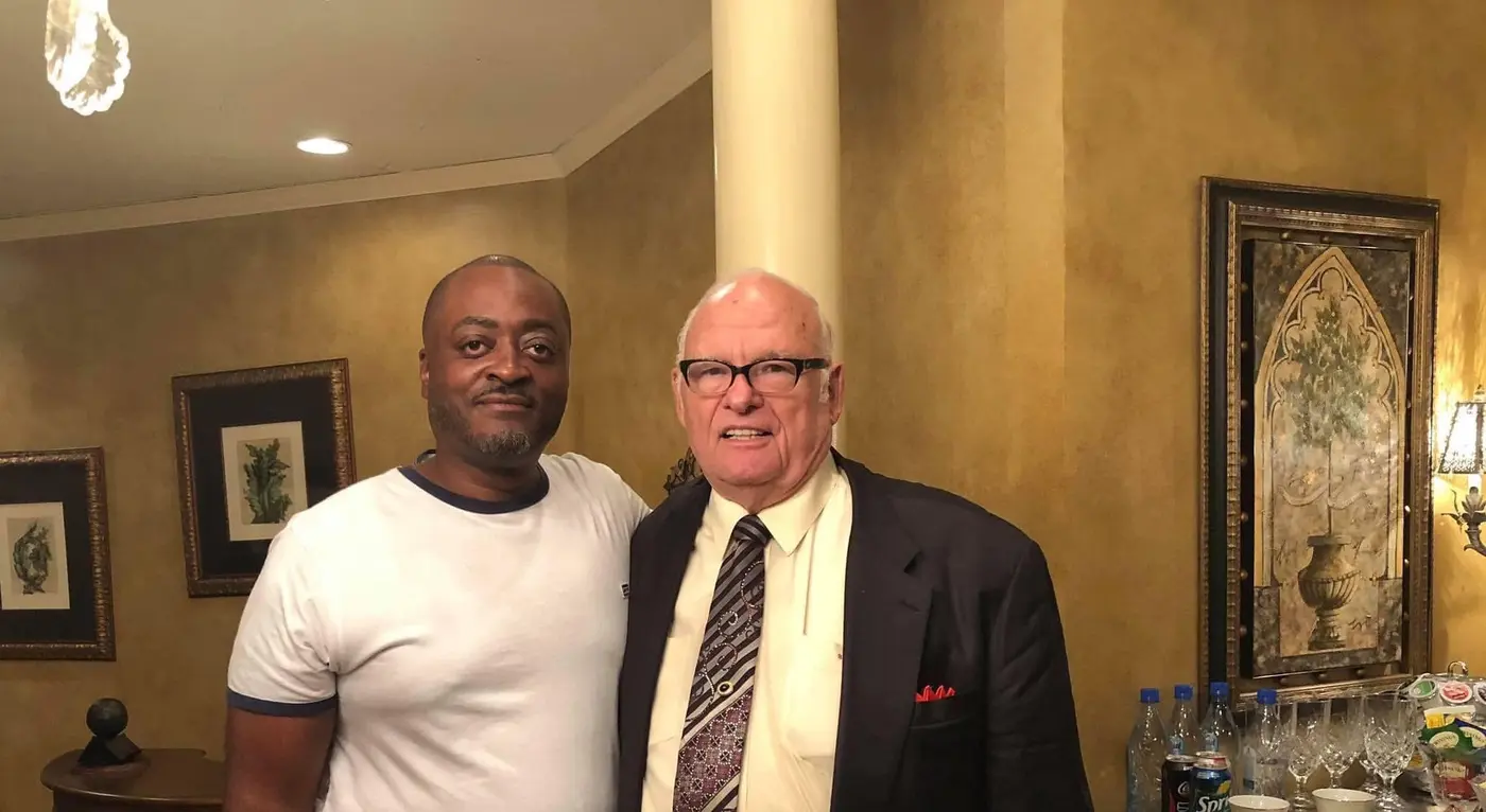 Melvin in a white t-shirt beside Apostle Eddie L. Long, in a dark jacket and patterned tie, in a room with gold walls