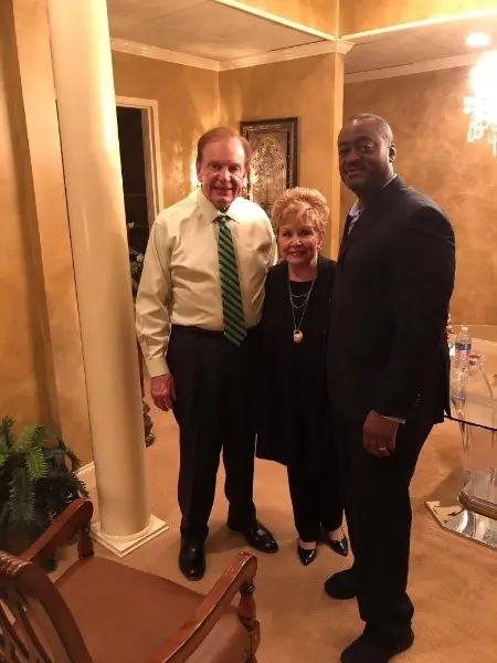 Melvin in a dark suit beside Apostle Buddy Crum, in a yellow shirt and green tie, and Dr. Mary Crum, in a room with gold walls and white columns