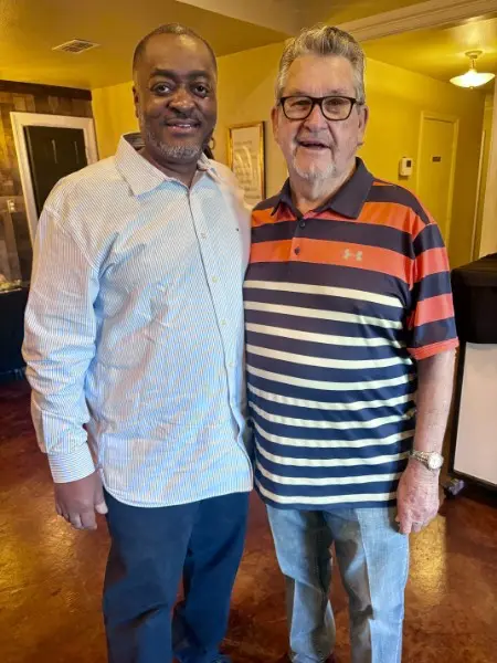 Melvin in a striped shirt beside Dr. Mark Hanby, an older man in glasses and a striped polo shirt, in a room with gold walls