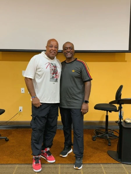 Melvin in a dark green t-shirt standing beside Bishop Vaughn McLaughlin, an older man in glasses, a white graphic t-shirt, and red sneakers, in a room with gold walls