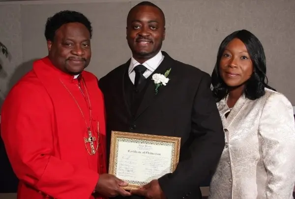 Melvin, in a dark suit with a white boutonniere, holding his framed Certificate of Ordination beside Apostle Eddie L. Long in red vestments and a woman in a cream jacket