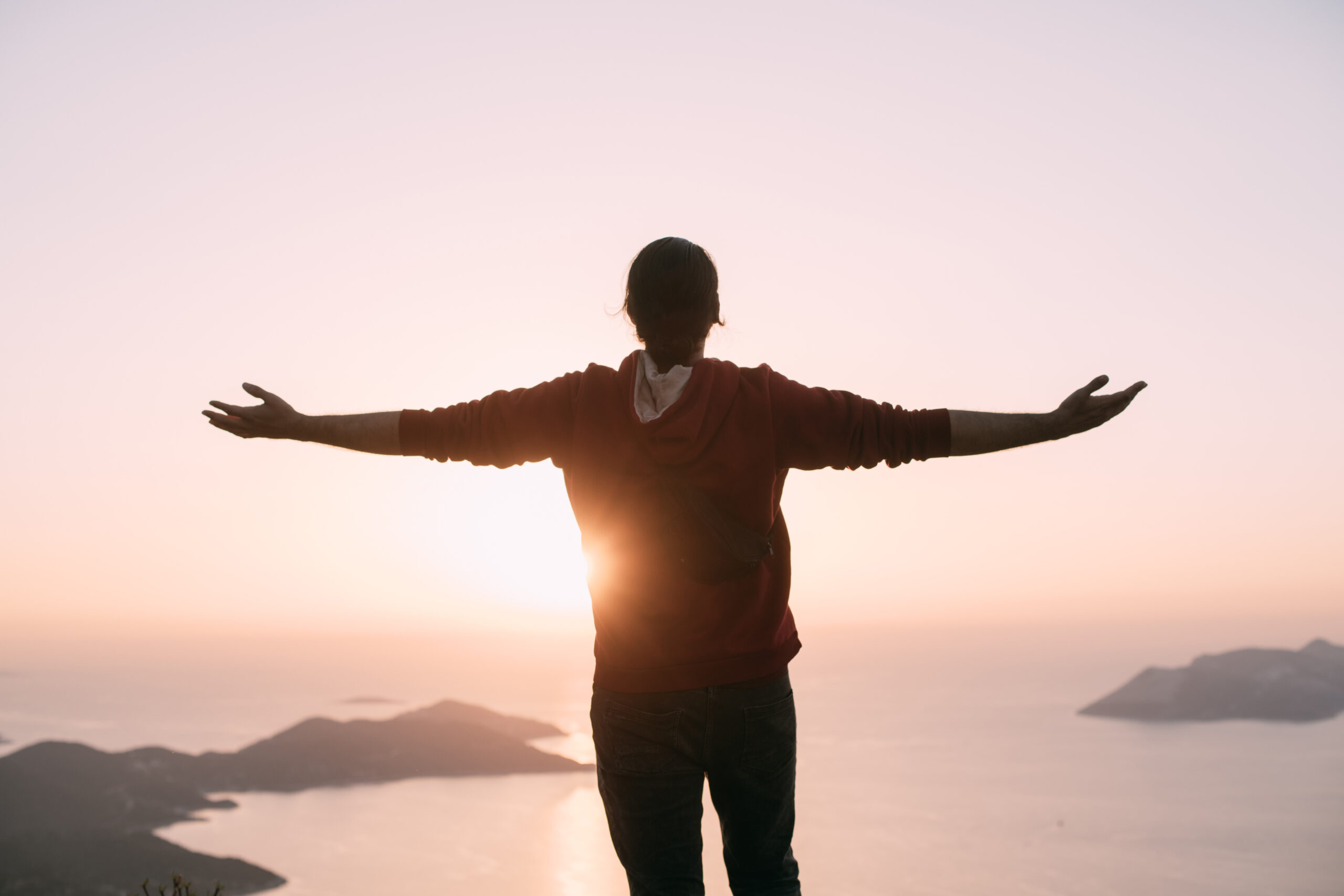 A young man meets the sunset, arms outstretched in a free gesture on a mountain by the sea. A Caucasian guy stands high on a cliff above a small town and looks at the sun setting behind the islands